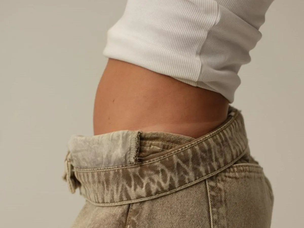 Woman wears white vest with grey jeans and shows tan lines on the pelvis on the subject of Tanrevel self-tanner