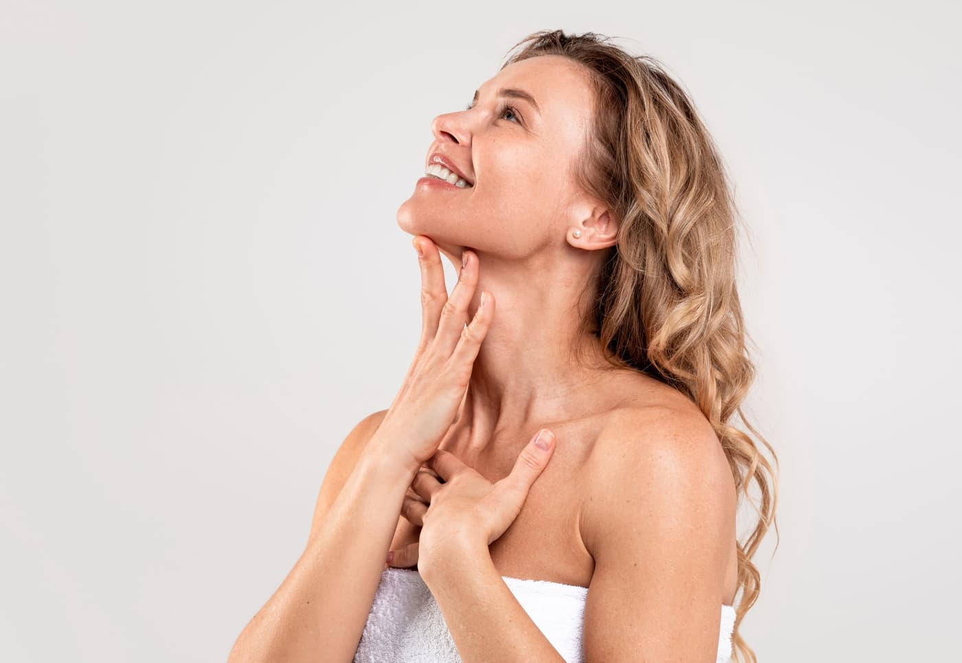 Smiling woman with sun-kissed skin gently applying moisturizing cream to her neck, bright studio background, fresh summer look.